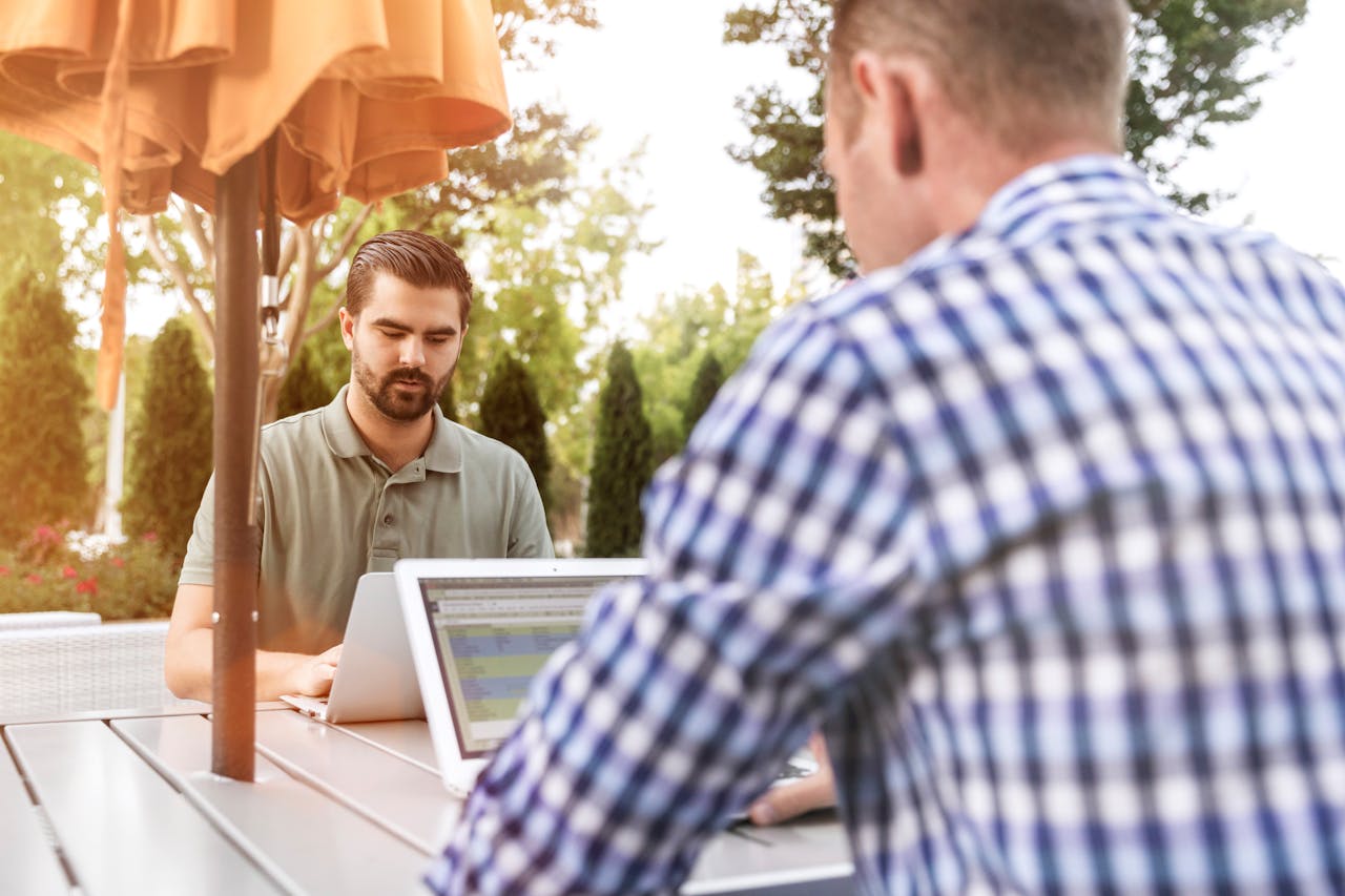 Men working on laptops outdoors, enjoying remote work and fresh air under a patio umbrella.